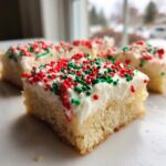 A close-up view of a square piece of christmas sugar cookie bars topped with thick white frosting and red and green holiday sprinkles.