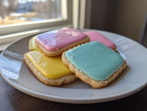 Four square sugar cookies topped with brightly colored, glossy sugar cookie icing in pink, yellow, and teal.
