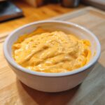 Close-up of thick, creamy, orange beer cheese dip served in a small white bowl on a wooden surface.