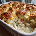A close-up of a creamy chicken pot casserole topped with golden brown biscuits, showing shredded chicken and vegetables.