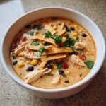 Close-up of a white bowl filled with creamy chicken tortilla soup, topped with crispy tortilla strips, black beans, corn, and cilantro.
