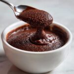 A spoonful of rich, creamy chocolate pudding being lifted from a white bowl, showing its thick texture.