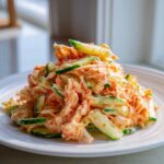 A mound of creamy kani salad featuring shredded imitation crab and sliced cucumbers served on a white plate.