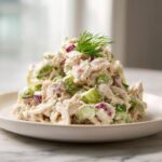 A close-up of a mound of creamy dill pickle chicken salad featuring shredded chicken, green pickle chunks, and red onion, topped with fresh dill.
