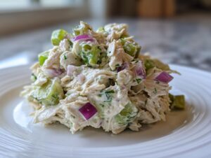 A mound of creamy dill pickle chicken salad featuring shredded chicken, bright green pickle chunks, and diced red onion on a white plate.