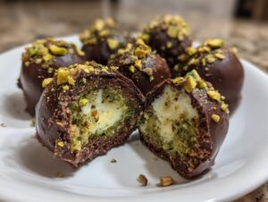 Close-up of Dubai chocolate balls, one cut open revealing a creamy white center surrounded by green pistachio filling, coated in dark chocolate and topped with chopped nuts.