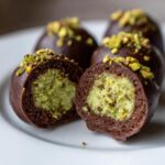 Close-up of a Dubai chocolate balls cut in half showing the bright green pistachio filling inside a dark chocolate shell, topped with chopped nuts.
