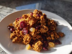 A mound of freshly baked gingerbread granola mixed with dried cranberries and candied ginger pieces, served on a white plate.