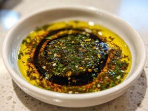Close-up of a white bowl containing olive oil bread dip, featuring bright green herbs, garlic, and a swirl of balsamic vinegar.