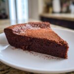 A decadent slice of flourless chocolate torte dusted with cocoa powder, served on a white plate.