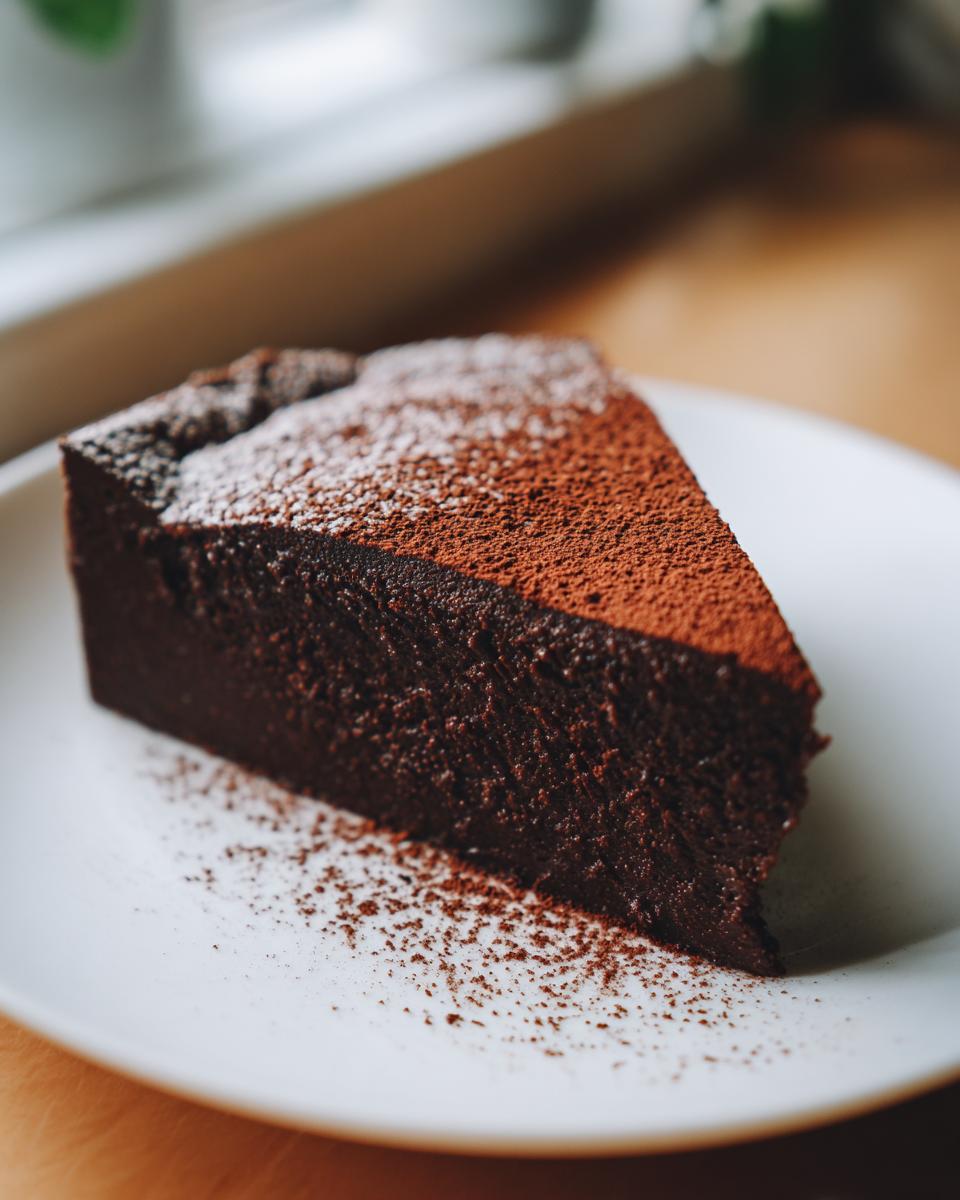 A dense, dark slice of flourless chocolate torte dusted with cocoa powder and powdered sugar on a white plate.