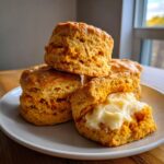 A stack of golden brown sweet potato biscuits, one split open and generously topped with cream cheese frosting.