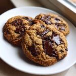 Close-up of three perfectly baked vegan chocolate chip cookies with melted chocolate chunks on a white plate.