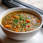 Close-up of a white bowl filled with hearty lentil soup, featuring visible lentils, diced carrots, and fresh parsley garnish.