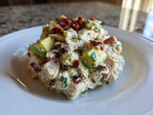 A scoop of creamy honey mustard chicken salad mixed with chunks of avocado and topped with crispy bacon bits, served on a white plate.