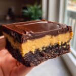 A close-up shot of a thick, three-layer buckeye brownies square held up, showing the brownie base, peanut butter layer, and chocolate topping.