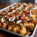 A close-up of fully loaded tater tot nachos covered in melted cheese, ground meat, bacon, diced tomatoes, and sour cream drizzle.