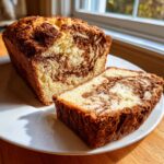 A freshly baked loaf of marbled cinnamon bread, with one thick slice cut and resting beside it on a white plate.