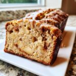 A close-up of a slice of moist apple bread, showing chunks of apple and a sweet glaze drizzled over the top.