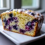Close-up of a moist slice of blueberry bread, packed with berries and topped with a sweet white glaze.