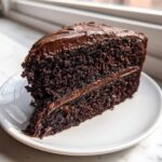 A thick slice of rich, dark chocolate cake recipe with creamy chocolate frosting, served on a white plate.