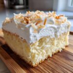 Close-up of a moist slice of coconut poke cake topped with whipped cream and toasted coconut flakes.