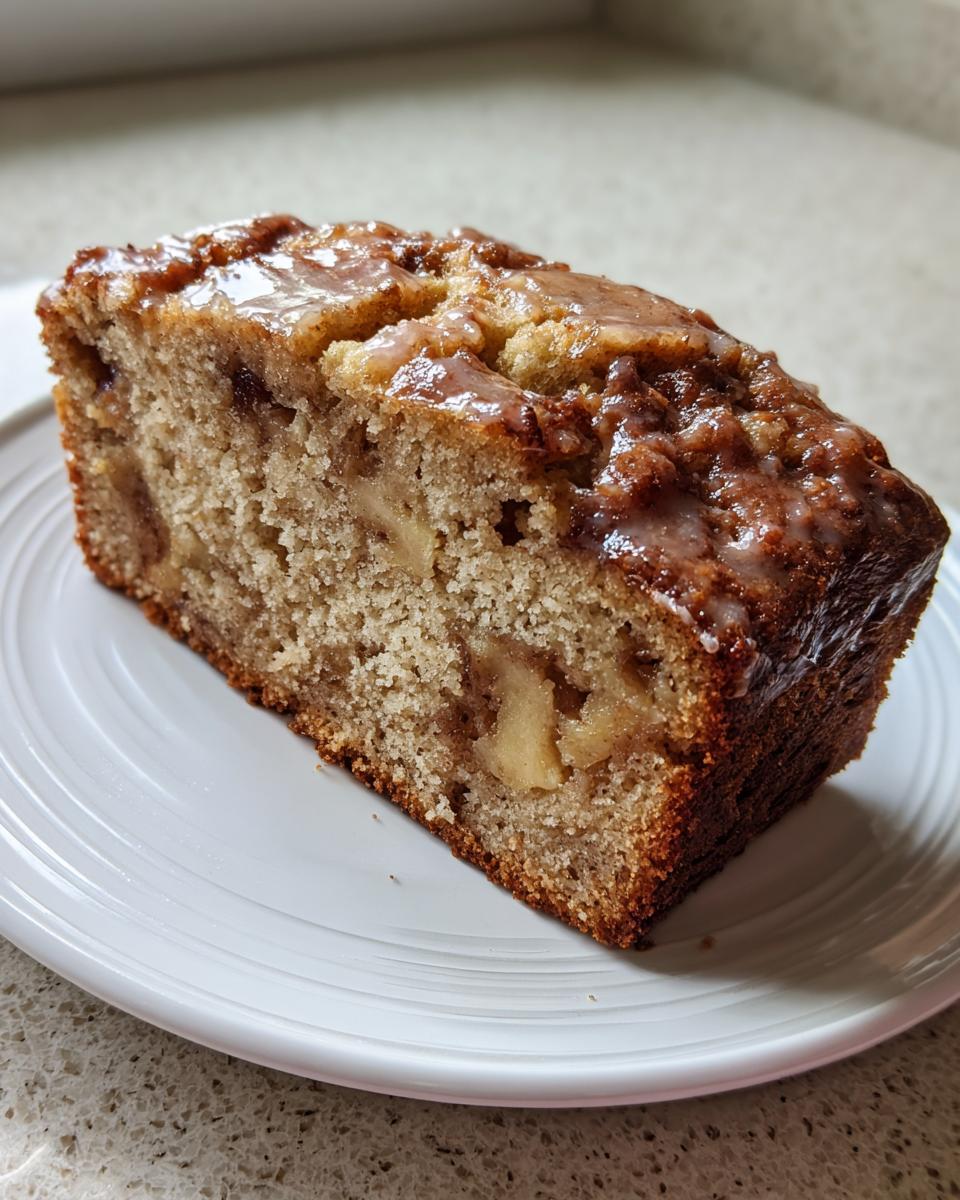 A close-up of a moist slice of apple bread with visible apple chunks and a shiny brown glaze on top.