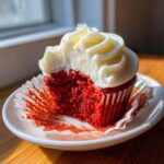 A bite taken out of a moist red velvet cupcakes showing the bright red interior and thick swirl of white cream cheese frosting.