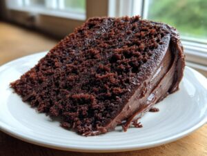 Close-up of a moist, rich slice of chocolate cake recipe with thick chocolate frosting on a white plate.