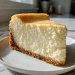 A thick, creamy slice of new york cheesecake with a golden-brown graham cracker crust resting on a white plate.