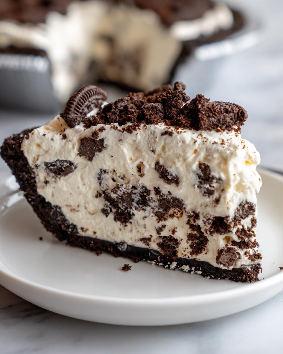 A close-up of a thick slice of no bake oreo pie featuring a dark crust, creamy filling studded with cookie pieces, and topped with crumbs.