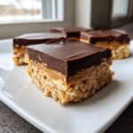 A close-up of a square no-bake peanut butter bars piece showing the crispy base, creamy peanut butter layer, and thick chocolate topping.