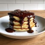 A tall stack of fluffy pancakes generously covered in rich, homemade chocolate syrup and chocolate shavings.