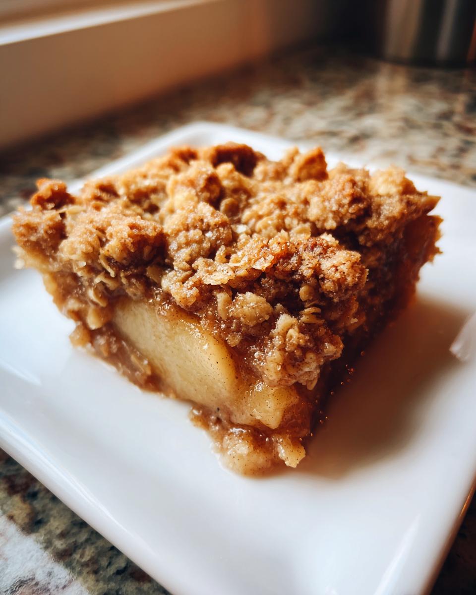 A close-up of a square slice of warm pear crisp showing soft spiced filling and a thick, crunchy oat topping.