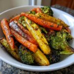 Close-up of vibrant roasted vegetables, including caramelized carrots and charred broccoli florets, seasoned in a white bowl.
