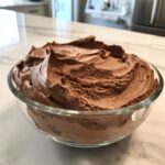A close-up of rich, airy chocolate mousse swirled high in a clear glass bowl.
