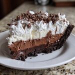 A rich slice of 8-layer mud pie featuring dark crust, chocolate filling, vanilla layer, and topped with whipped cream and chocolate shavings.