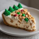 A close-up slice of Christmas tree cheesecake on a white plate, topped with green frosting trees and candy.