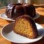 Close-up of a moist slice of rum cake recipe with rich chocolate glaze, whole cake in background.