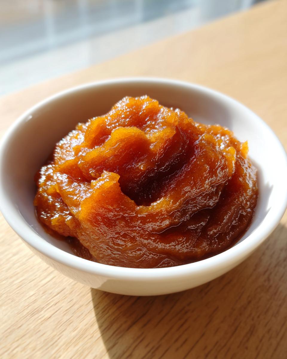 A close-up of thick, glossy, dark orange pear butter served in a small white bowl on a wooden surface.