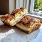 Two halves of a rich sopapilla cheesecake showing flaky pastry and creamy filling, dusted with cinnamon sugar.
