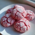 Five bright pink strawberry cake mix cookies covered in cracked powdered sugar on a white plate.