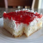 A close-up of a moist slice of strawberry jello poke cake topped with white whipped cream and bright red strawberry gelatin.