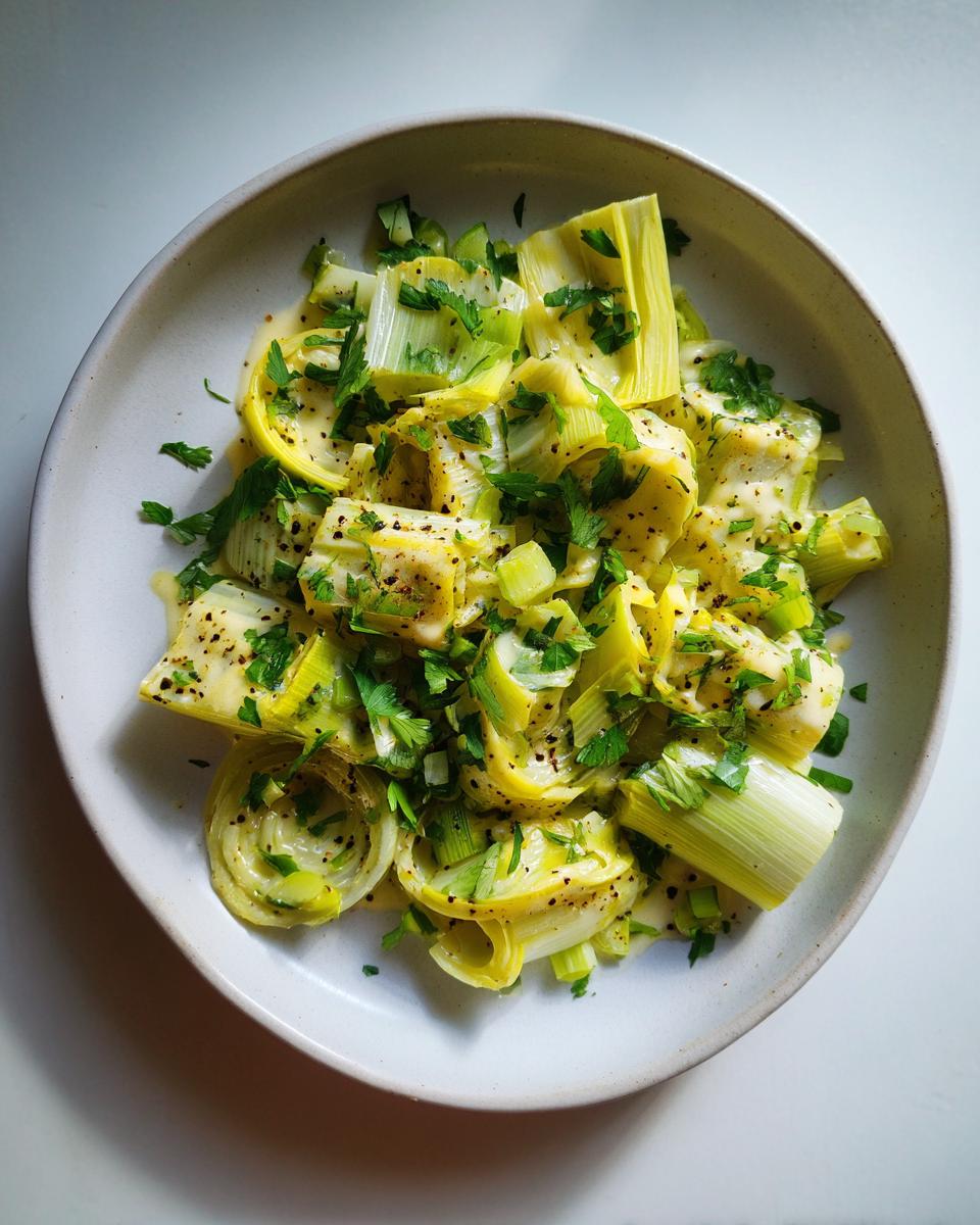 Overhead view of tender braised leeks served in a light sauce, topped generously with fresh parsley and black pepper.