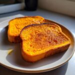 Two slices of bright orange sweet potato toast, lightly toasted and drizzled with syrup, sitting on a light gray plate.