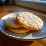 Three golden vegan sugar cookies stacked on a light grey plate, generously topped with sparkling sugar crystals.