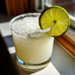 A frosty glass of a white Christmas margarita with a salt rim and a bright lime wheel garnish.