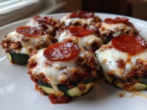 Six baked zucchini pizza bites topped with melted mozzarella, sauce, and pepperoni slices on a white plate.