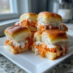 A stack of freshly baked buffalo chicken sliders oozing with orange sauce and white ranch dressing on a white platter.