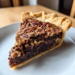 Close-up of a rich slice of chocolate pecan pie showing the flaky crust and gooey filling topped with pecans.